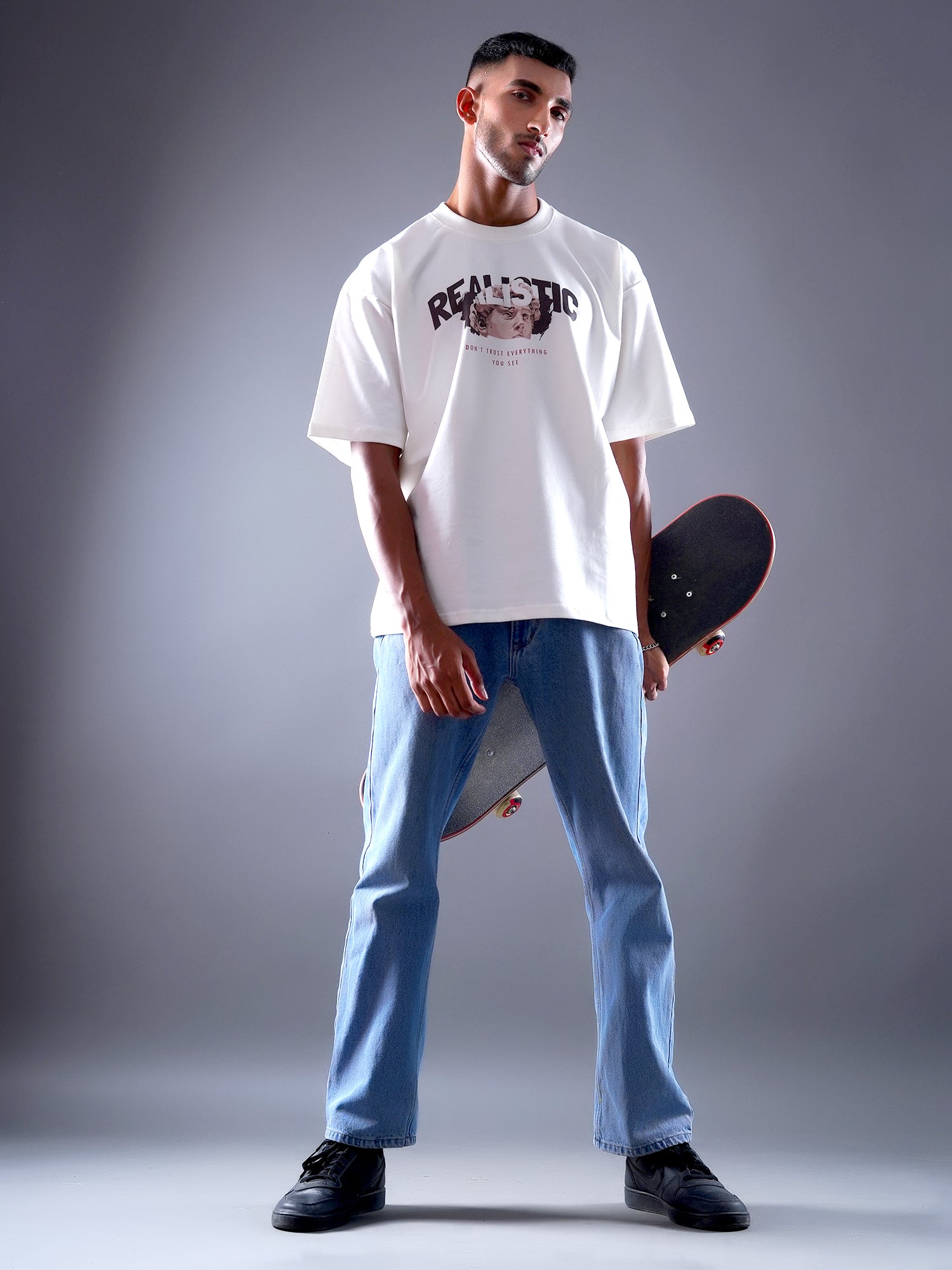 Man holding a skateboard wearing a white t-shirt and blue jeans on a gray background